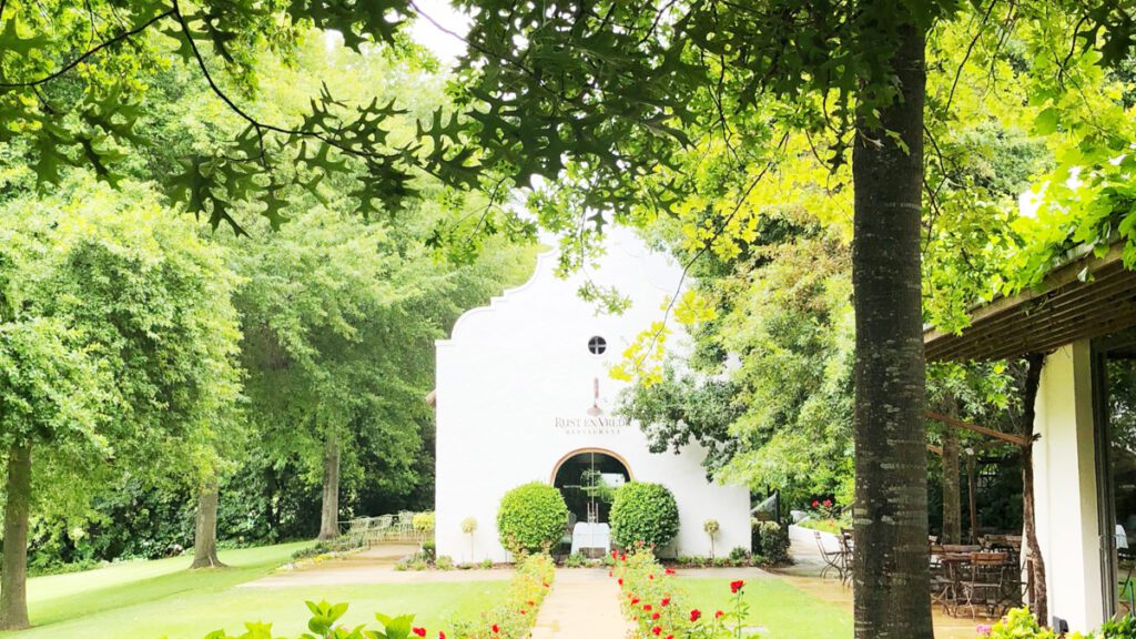 En typisk Cape Dutch-byggnad i vitt skymtar bakom en massa grönt - träd, blommor, gräsmatta. Rust En Vrede är vår absoluta favoritrestaurang i Stellenbosch.